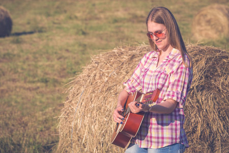 Young fashionable woman with acoustic guitar leaning against hay roll outdoorsの写真素材