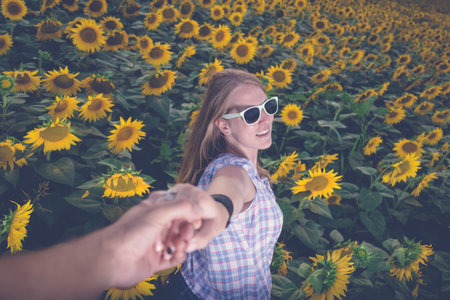 Young modern woman in sunglasses holding man's hand in sunflower field. Summertime and love concept.の写真素材