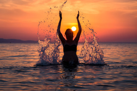 Beautiful female in ocean splashing water with hands. Sunset in the background. Nature and summer...