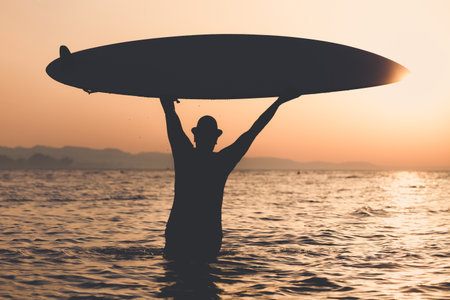 Tourist guy in hat standing in water and holding up surfboard at sunset. Freedom and summer vacation concepts.の写真素材