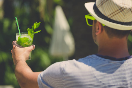 Rear view of man in hat and sunglasses holding mojito cocktail. Summer vacation concept.の写真素材