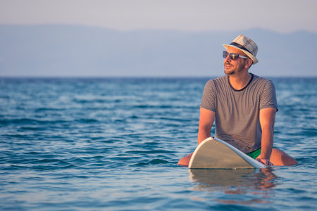 Man in hat and sunglasses sitting on surfboard in ocean. Summer vacation and travel concepts.の写真素材