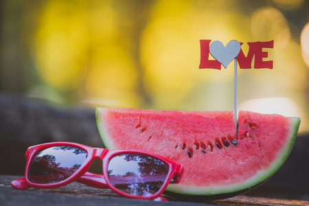 Close up of watermelon slice with love sign on stick and sunglasses on table in the park. Healthy eating and summertime concepts.の写真素材