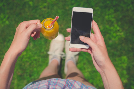 High angle view of female holding glass bottle of fresh orange juice and blank screen smartphone on field. Healthy lifestyle and technology concepts. の写真素材