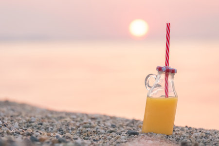 Glass of orange juice with drinking straw on beach. Sunset and ocean in the background. Healthy eating and summer vacation concepts. Space for copy.の写真素材