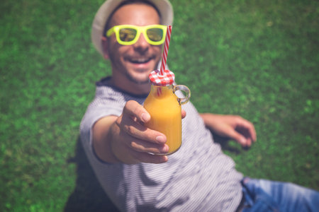 Portrait of young guy lying on the field and showing a glass bottle of fresh orange juice. Summertime and healthy lifestyle concepts.の写真素材