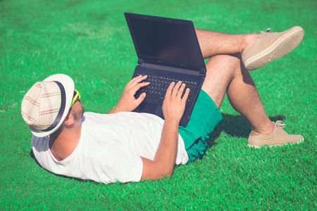 Young guy in hat and sunglasses lying on field and using blank screen laptop. Summertime and technology concepts.の写真素材