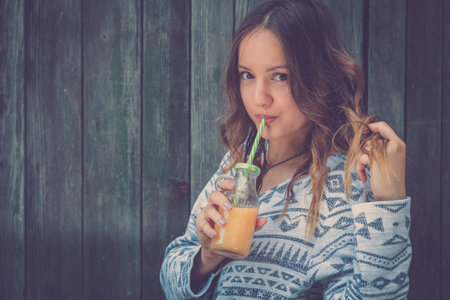 Portrait of young fashionable woman drinking orange smoothie against old wooden wall outdoors. Healthy lifestyle and fashion concepts.の写真素材