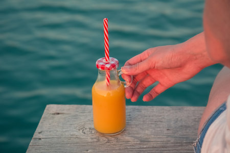 Close up of female hand holding glass bottle of fresh orange juice with drinking straw at the edge of wooden pier. Sea in the background. Summertime and healthy eating concepts. の写真素材