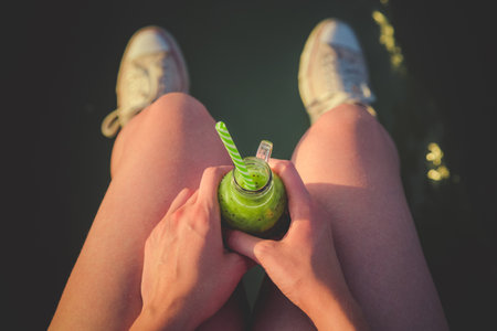 High angle view close up of young female holding glass bottle of fresh green kiwi smoothie with drinking straw between her knees against water surface background. Summertime and healthy lifestyle concepts.の写真素材
