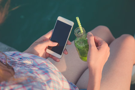 High angle view of female holding glass bottle of fresh green kiwi juice and blank screen smartphone against water surface background. Healthy lifestyle and technology concepts.の写真素材