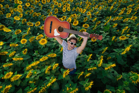 Portrait of young modern guy holding acoustic guitar in sunflower fieldの写真素材