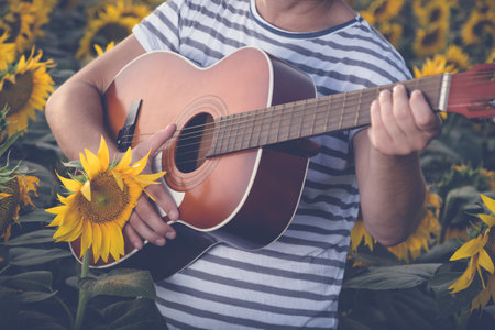 Close up of young modern guy playing acoustic guitar in sunflower field. Music and nature concepts.の写真素材