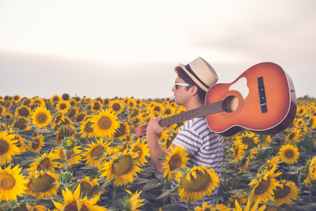 Young retro styled man with acoustic guitar over his shoulder walking through the sunflower field. Sun and clouds in the background. Music, art and lifestyle concepts.の写真素材