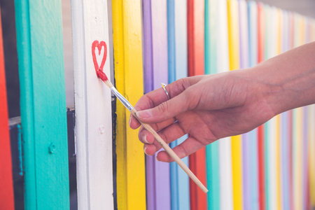 Close up of female hand holding paint brush and drawing red heart shape on colorful wooden fence outdoor. Love and art concepts.の写真素材
