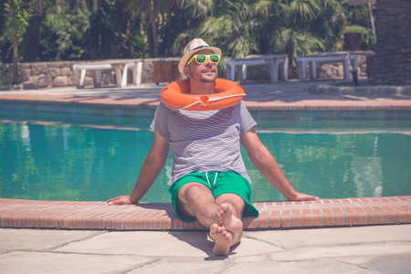 Portrait of young guy with hat, sunglasses and lifebuoy relaxing next to the swimming pool. Summer...