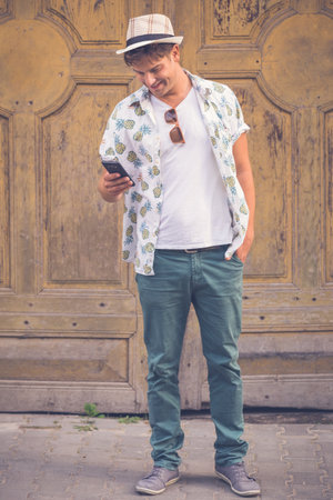 Cheerful hipster guy holding and looking at smart phone against old retro door backgroundの写真素材