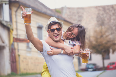 Cheerful young hipster couple holding mugs with beer walking on the city streetの写真素材