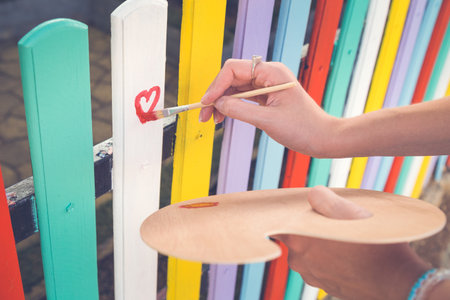 Close up of female hand holding paint brush and drawing red heart shape on colorful wooden fence...