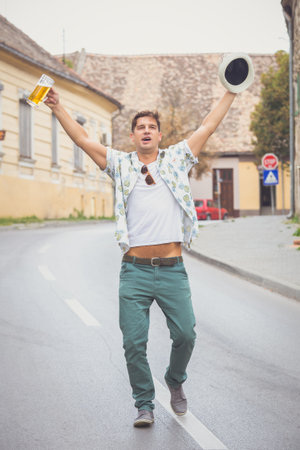 Portrait of cheerful drunk guy walking on the street and singing with hands up. He is holding stein full of beer and hat. の写真素材
