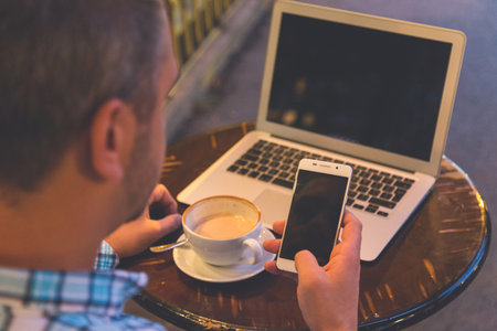 Businessman using blank screen smart phone and laptop on wooden table with cup of latte macchiato in...