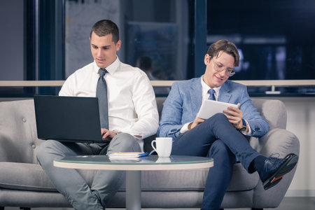 Two young businessmen sitting in office lobby and using laptop and digital tablet deviceの写真素材
