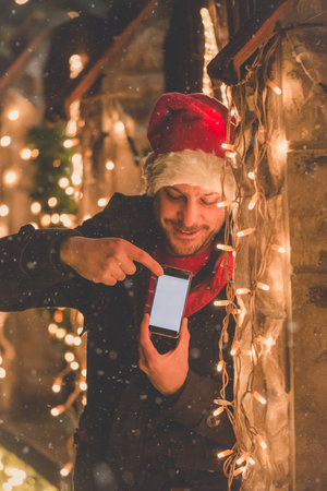 Portrait of young man with Christmas hat standing next to Christmas lights decoration and showing blank screen smartphone. Space for copy.の写真素材