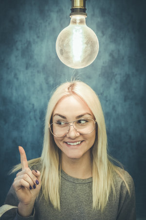 Happy thoughtful woman with light bulb above head isolated on blue wall background. Thinking and idea concepts.の写真素材