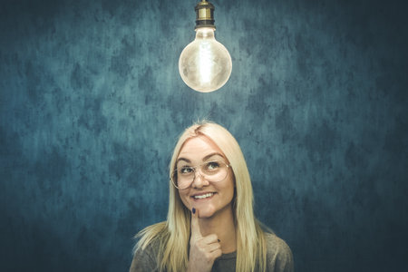 Happy thoughtful woman with light bulb above head isolated on blue wall background. Thinking and idea concepts.の写真素材