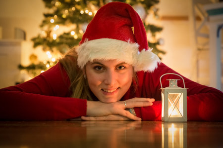 Portrait of beautiful girl in santa hat lying on floor next to lantern. Decorated christmas tree in the background. Holiday concept.の写真素材