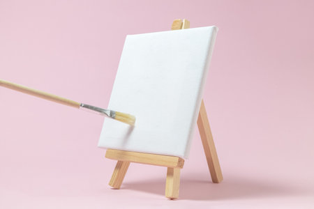 Blank art canvas on easel and paintbrush against pastel pink rose background. Artist minimal concept. Space for copy.の写真素材