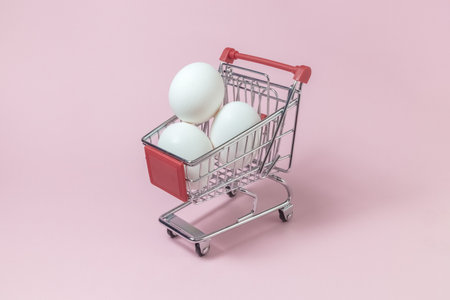 Shopping trolley model full of white eggs against pastel pink rose background minimal concept. Space for copy.の写真素材