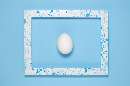 Flat lay of white egg in wooden frame colored with blue paint splatters on blue background minimal concept.の写真素材