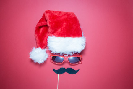 Flat lay of Santa hat, red sunglasses and mustache paper props in form of hipster Christmas guy on red background minimal concept.の写真素材