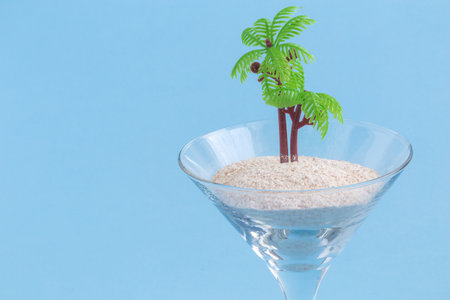 Crystal cocktail glass with sand and palm tree on pastel blue background. Summer minimal concept.の写真素材
