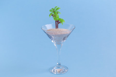 Crystal cocktail glass with sand and palm tree on pastel blue background. Summer minimal concept.