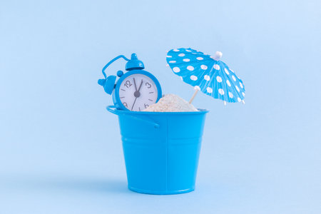 Alarm clock, small bucket full of sand and paper umbrella against pastel blue background. Summertime minimal conceptの写真素材