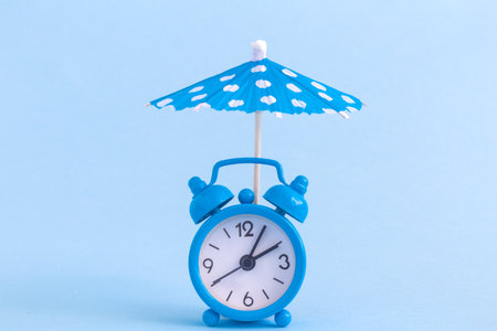 Alarm clock and paper umbrella on pastel blue background. Summer vacation minimal concept.