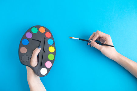 Female hands holding art palette with paints and paintbrush against blue background minimal creative concept.の写真素材