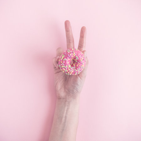 Close up of female hand holding small donut and showing victory sign with fingers on pastel pink background minimal creative concept.の写真素材