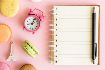 Flat lay of colorful macaroons, small alarm clock, Eiffel Tower and clean notebook against pastel pink background.の写真素材