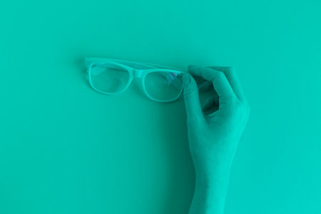 Female hand holding eyeglasses. Turquoise colored minimal creative concept.の写真素材