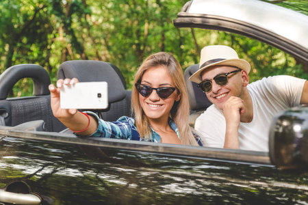 Young couple taking selfie with smartphone while sitting in convertible car. Travel and technology concepts.の写真素材