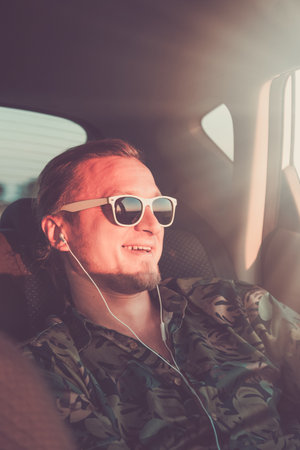 Portrait of young hipster guy with earphones sitting on back seat. Travel and music concepts.