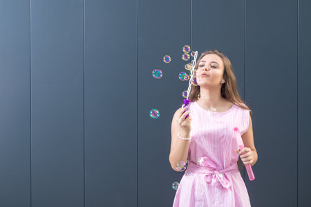 Beautiful young girl blowing soap bubbles against gray background. Space for copy.