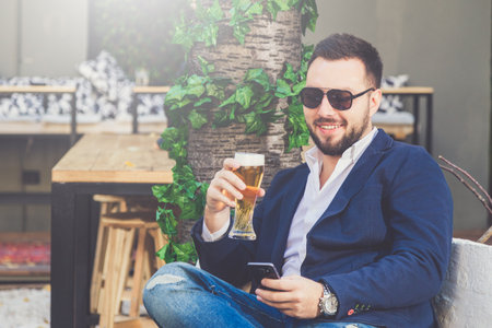 Modern fashionable guy using mobile phone and drinking beer in cafe lounge.の写真素材