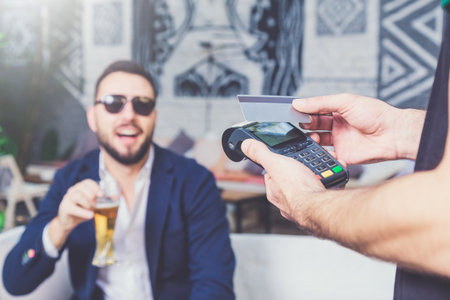 Paying with credit card in cafe. Businessman holding glass of beer in the background.の写真素材