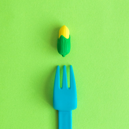 Flat lay of plastic fork and small rubber corn toy against green background minimal creative vegetarian concept.の写真素材