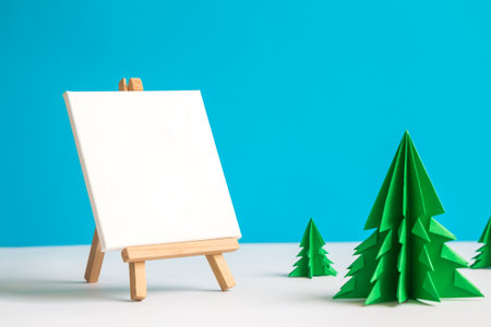 Easel for artists and blank canvas with christmas trees made of paper against pastel blue background.の写真素材