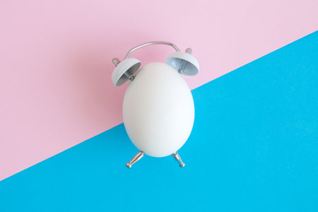 Flat lay of white egg in form of alarm clock on pastel background minimal easter time creative concept.の写真素材